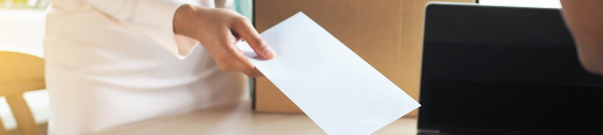 Business woman has a brown cardboard box next to her and hands a letter of resignation to a seated businessman sitting at his desk. 